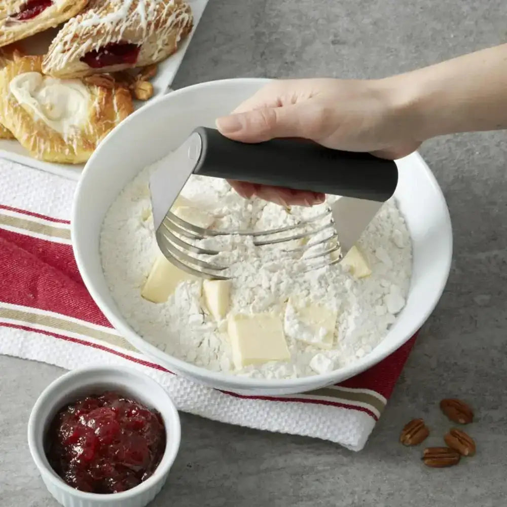 Person using a pastry blender to blend butter into flour in a bowl, with pastries and jam in the background.