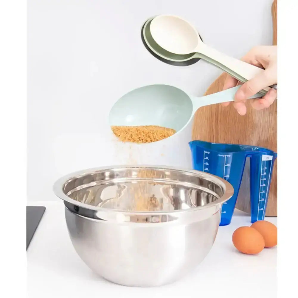 Person using a measuring spoon to add ingredients into a metal mixing bowl on a white surface.