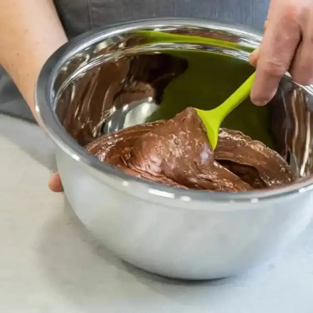 Person using a green spatula to mix chocolate in a metal bowl.