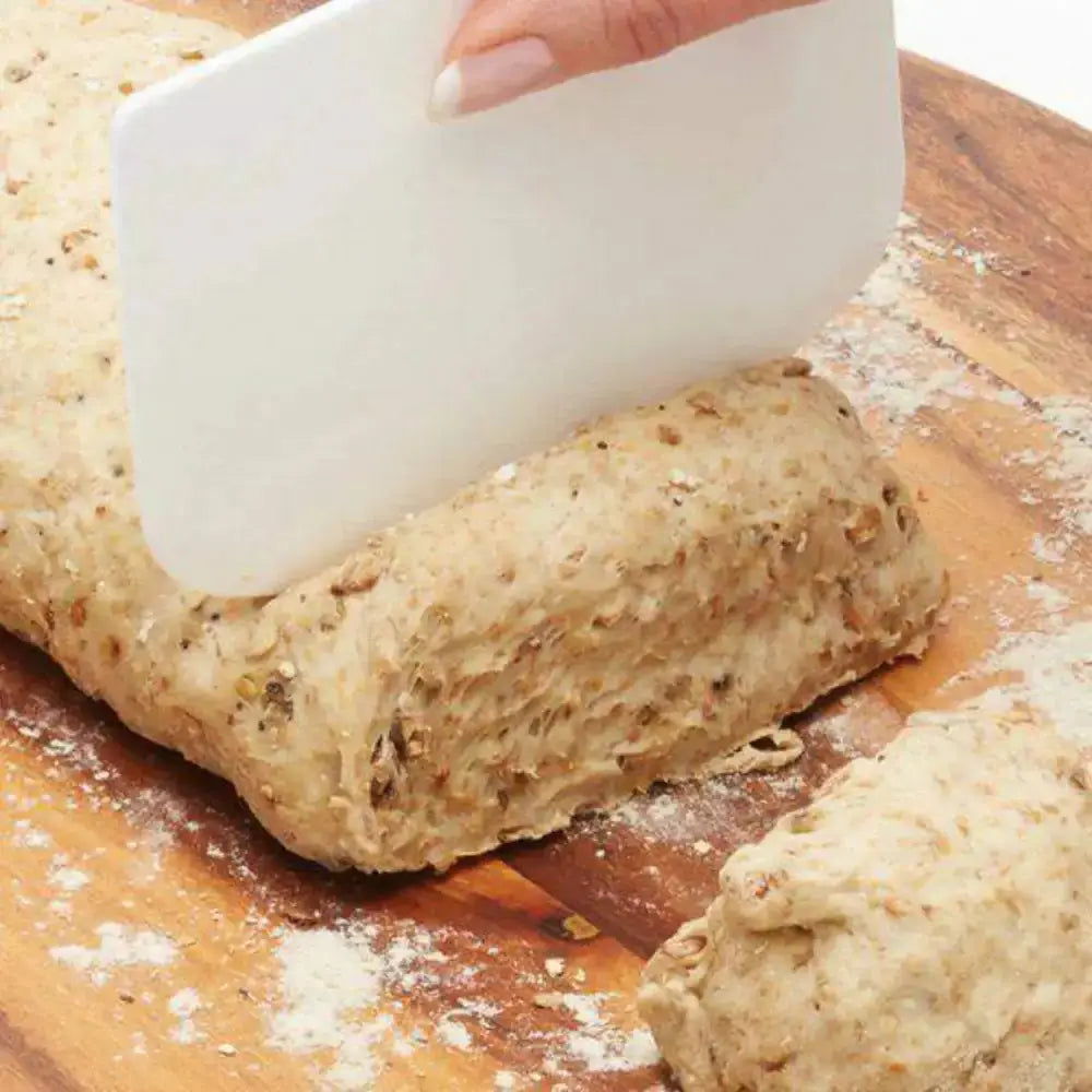 Hand using a white plastic scraper to cut through a loaf of bread on a wooden cutting board.