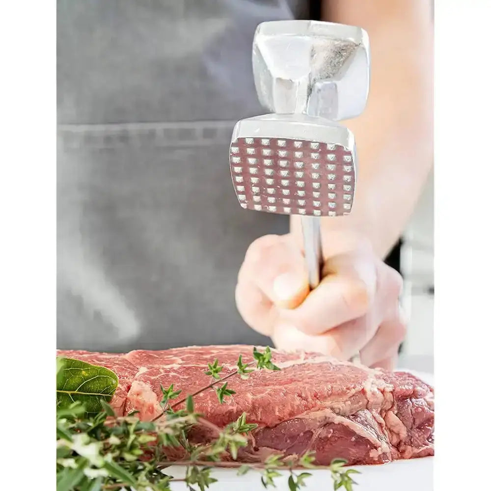 Hand using a meat tenderizer on a piece of raw meat with herbs.