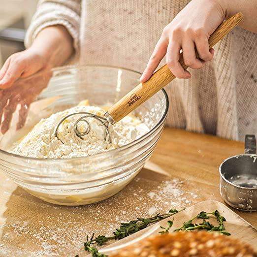 Person using a wooden dough scraper in a bowl of flour on a wooden table.