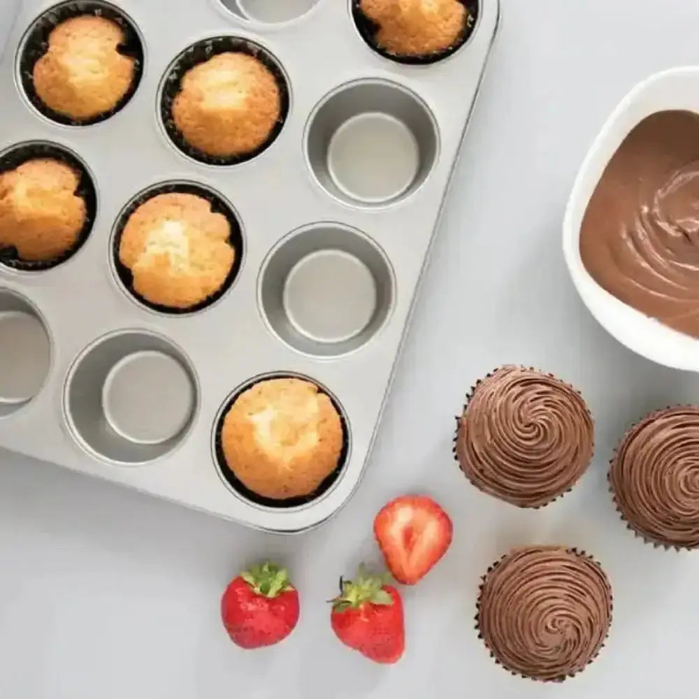 Muffin tin with baked muffins, a bowl of chocolate frosting, and frosted cupcakes on a light surface.