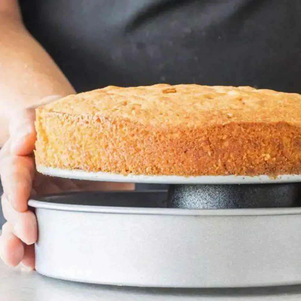 Baked cake in a round metal baking pan held by a person wearing gloves.