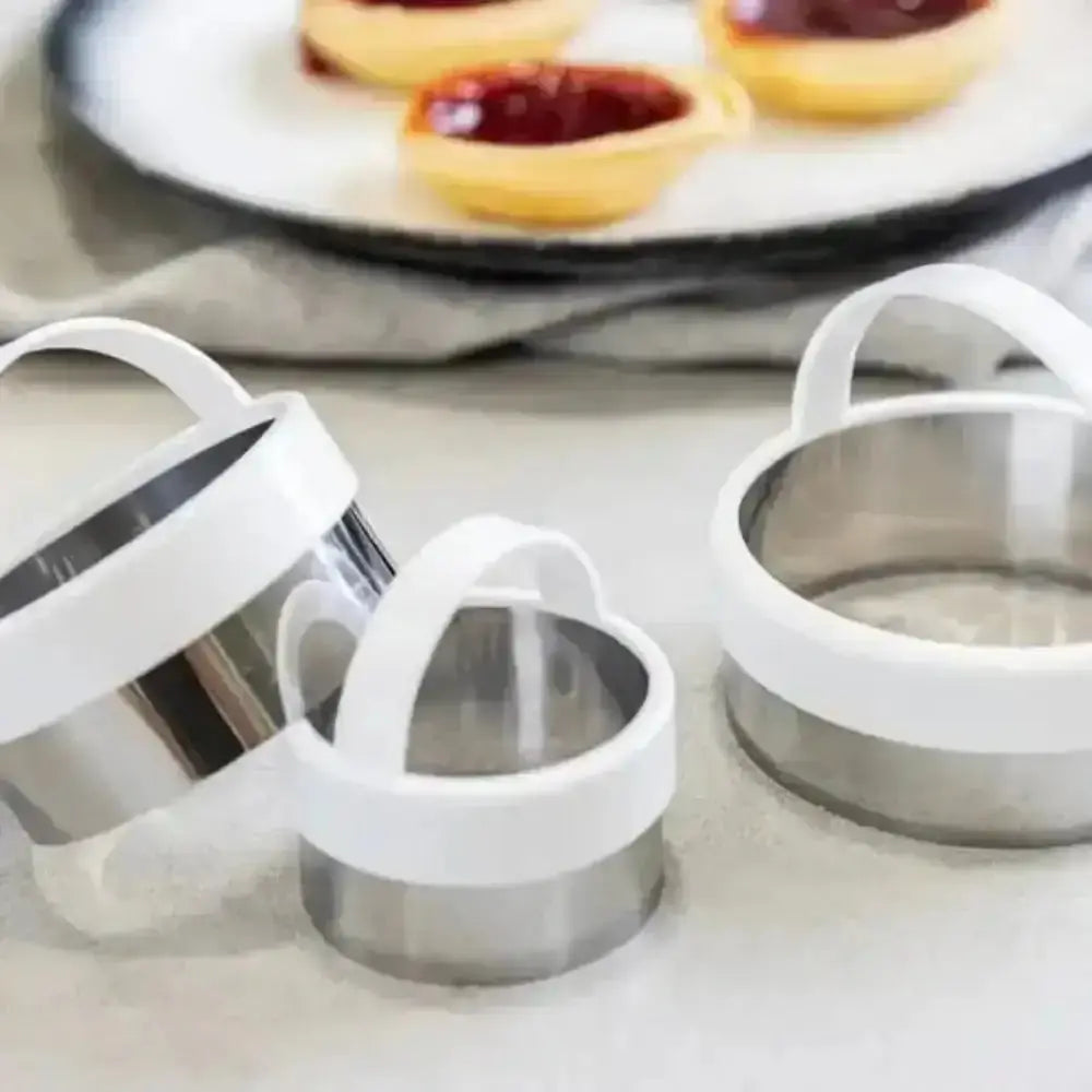 Metal cookie cutters with white handles on a light surface, with pastries in the background.