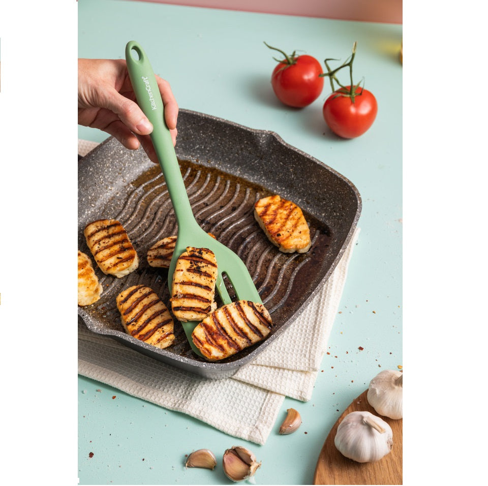 Person using a green spatula to serve roasted vegetables from a cast iron pan on a light blue surface.