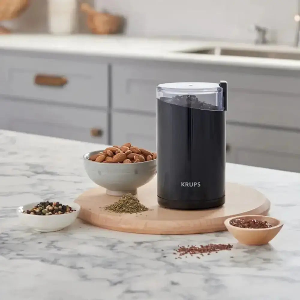 Black KRUPS coffee grinder on a kitchen counter with bowls of spices and nuts.