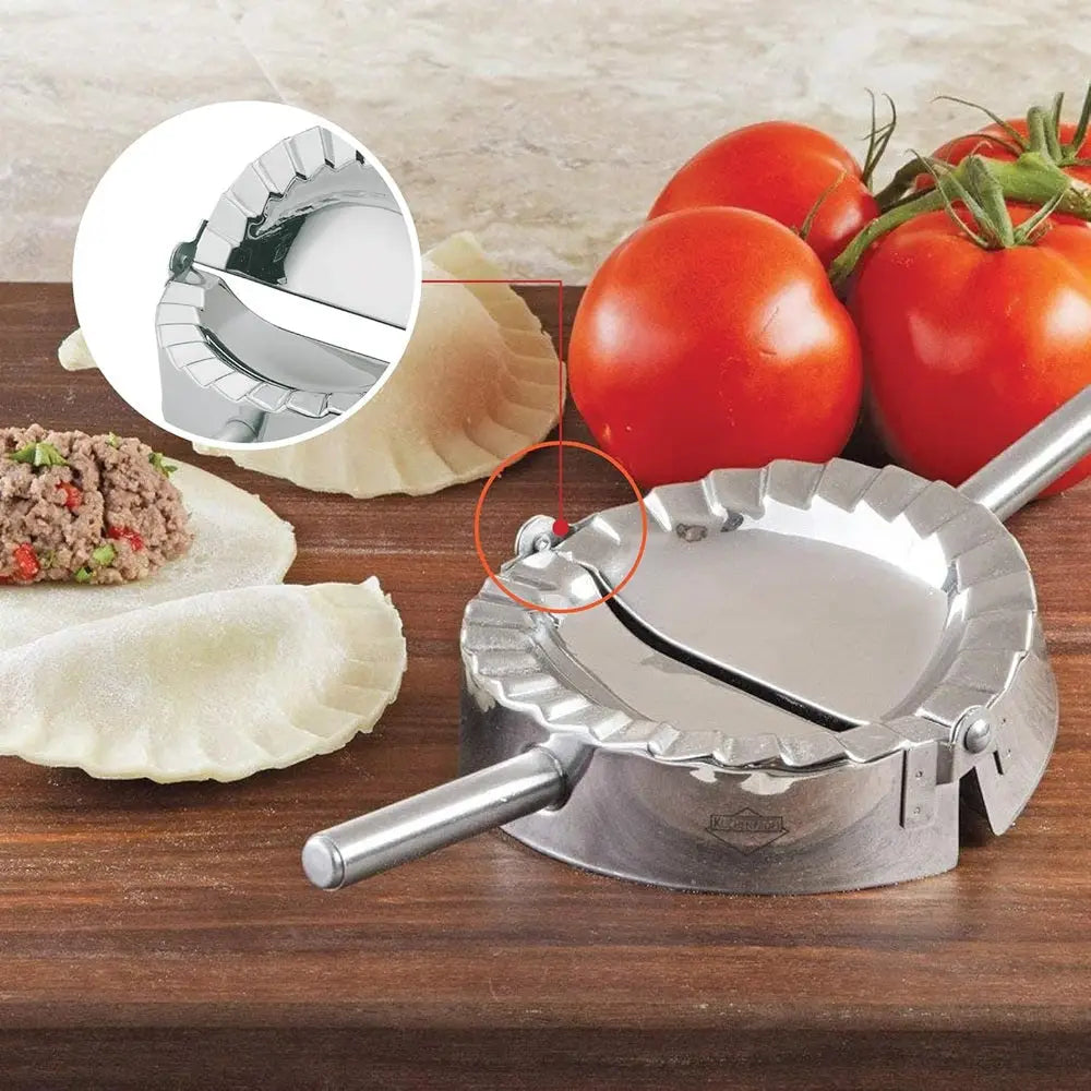 Metal dumpling maker on a wooden surface with tomatoes and dumplings.