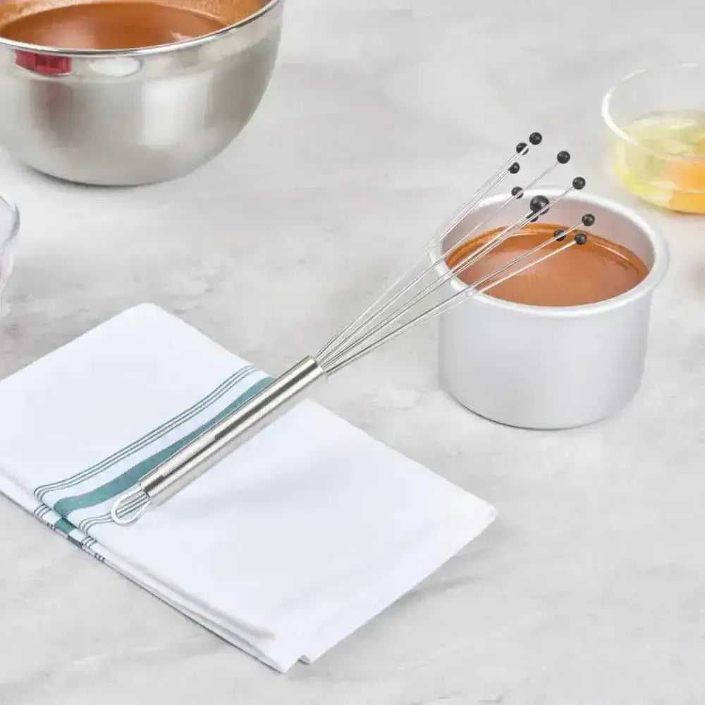 Metal whisk on a white napkin with a small pot of brown liquid and a bowl in the background.