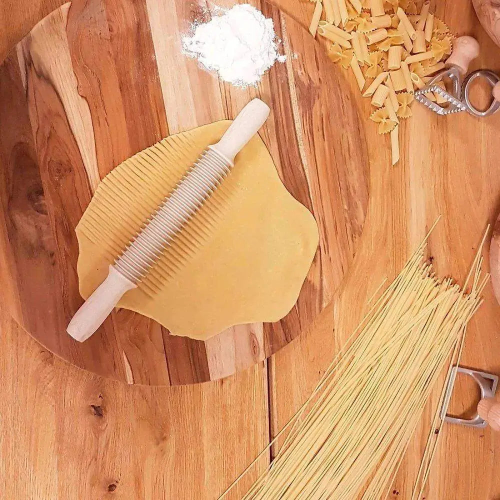 Wooden cutting board with pasta, a rolling pin, and a pasta shaper on a wooden surface.