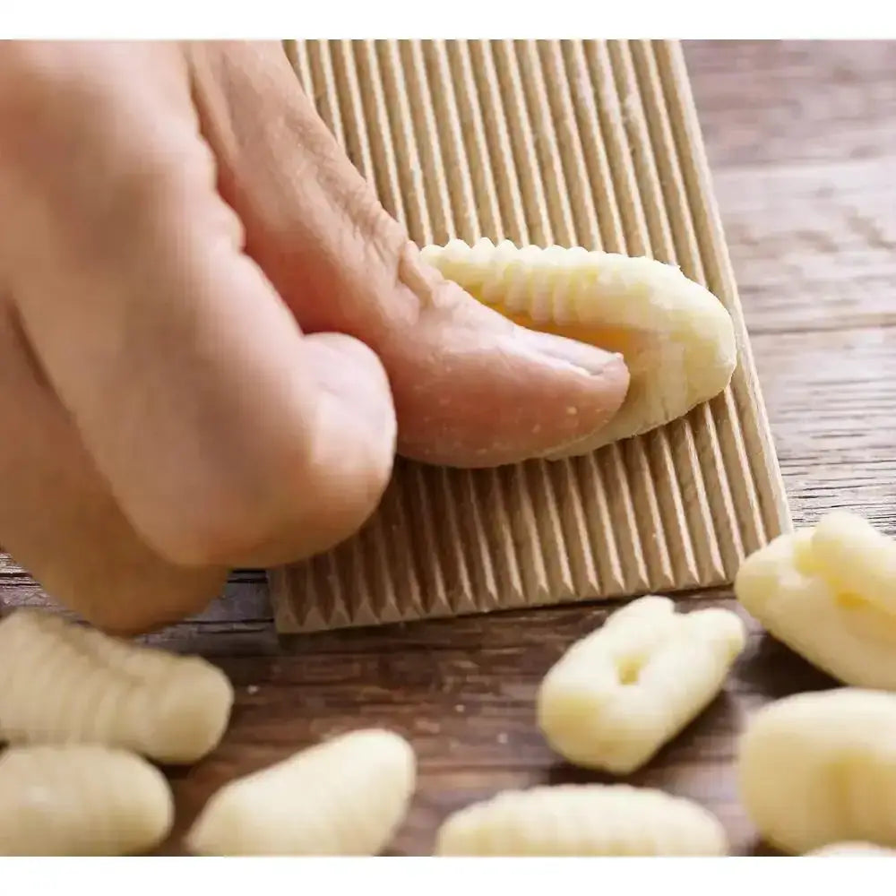 Hand rolling a piece of pasta on a wooden surface with other uncooked pasta pieces around.