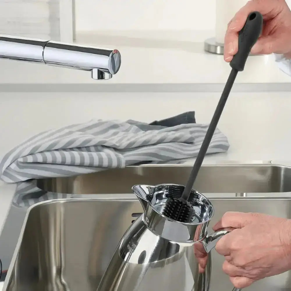 Person cleaning a stainless steel pitcher with a brush over a sink.