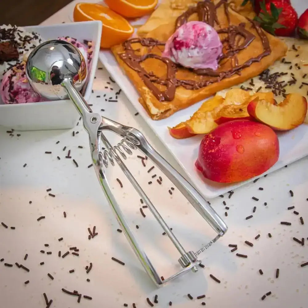 Silver ice cream scoop on a table with dessert items including waffles, fruit, and ice cream.