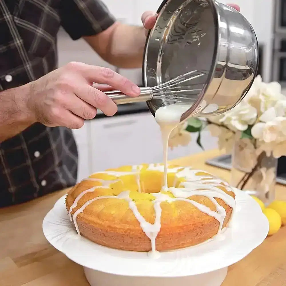 Person pouring a white glaze over a cake with a whisk on a kitchen counter.