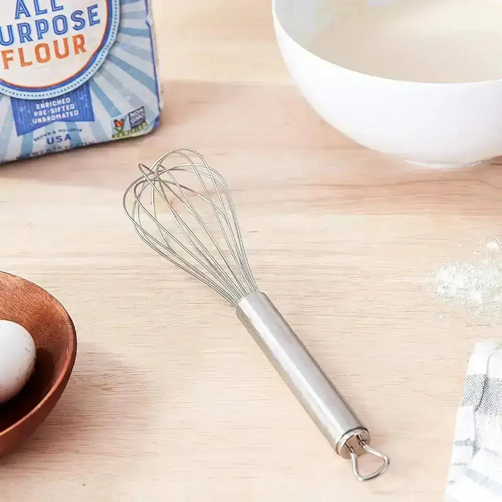 Metal whisk on a wooden surface with flour and an egg in the background