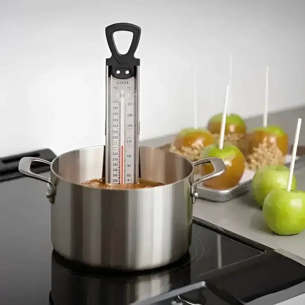 Stainless steel pot with candy thermometer on a stove, caramel apples in the background