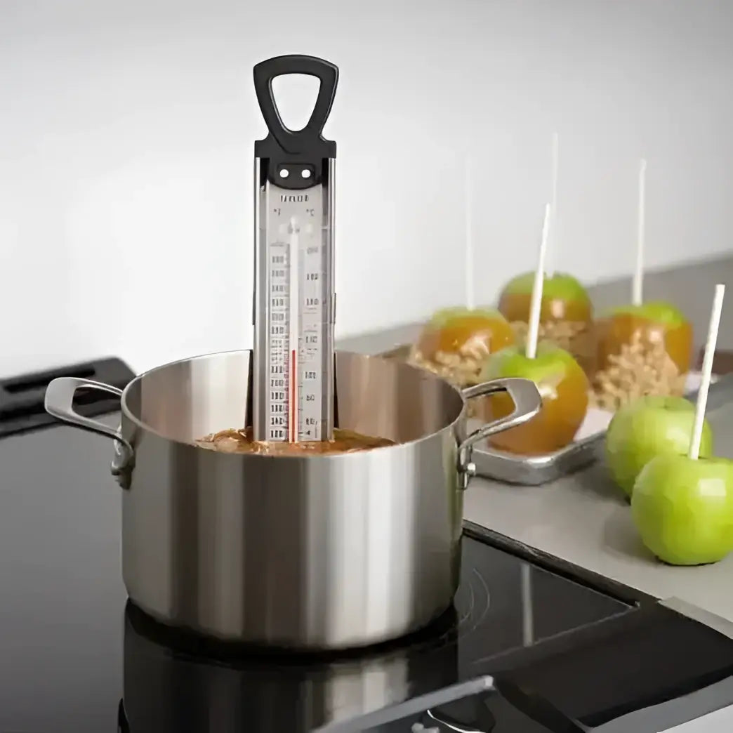 Stainless steel pot with candy thermometer on a stove, caramel apples in the background