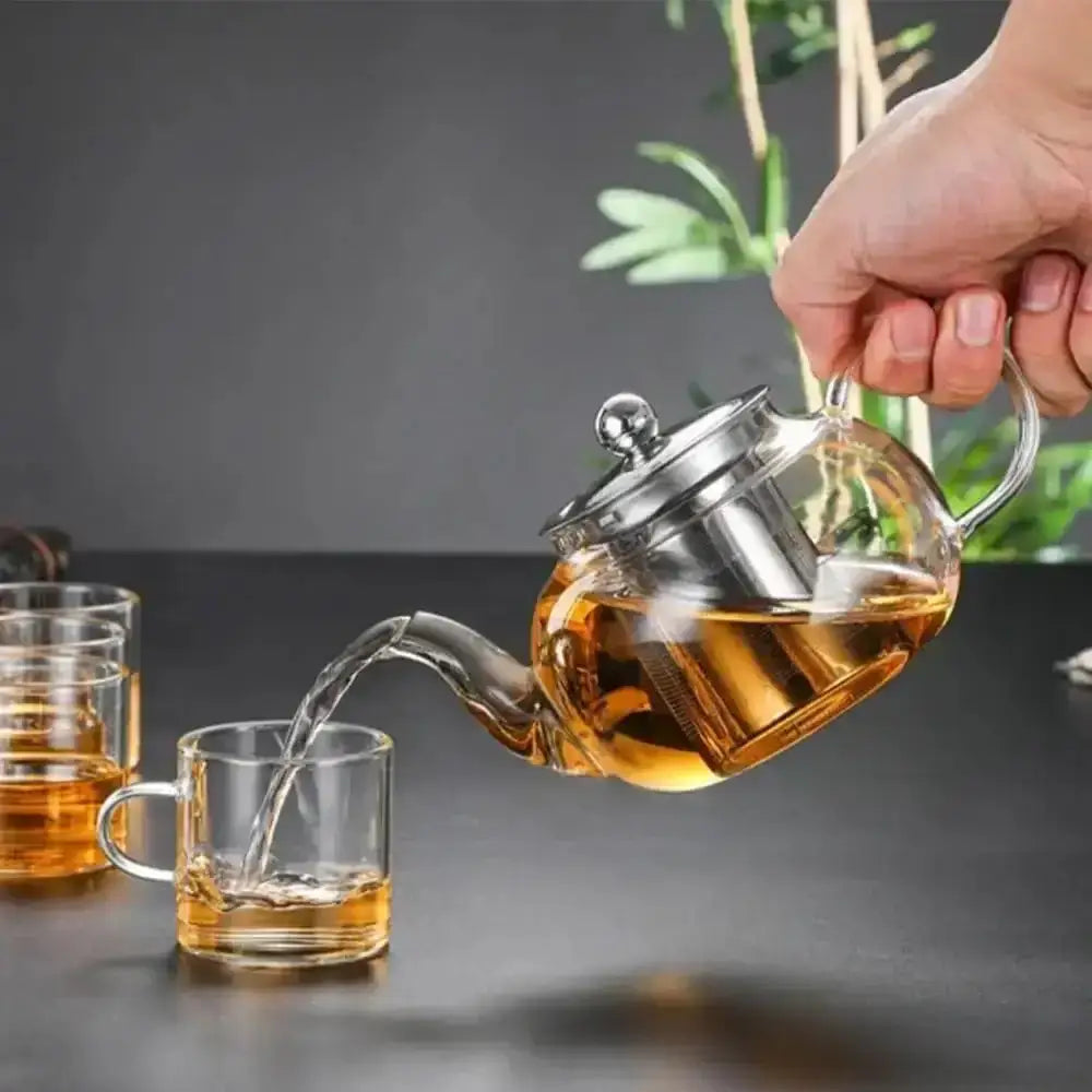 Tea being poured from a glass teapot into a clear mug on a dark surface with a blurred plant in the background.
