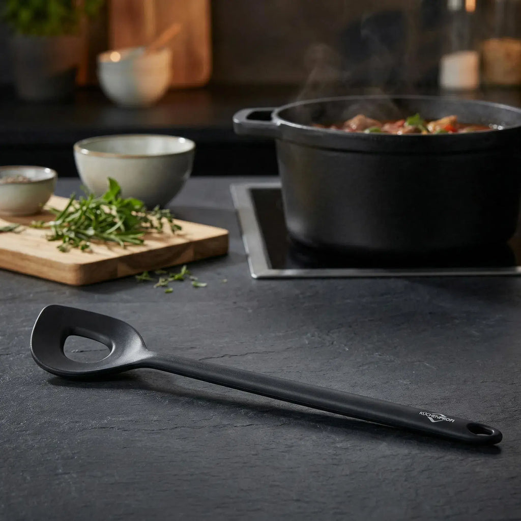 Black mixing spoon on a kitchen counter with a pot of food and ingredients in the background