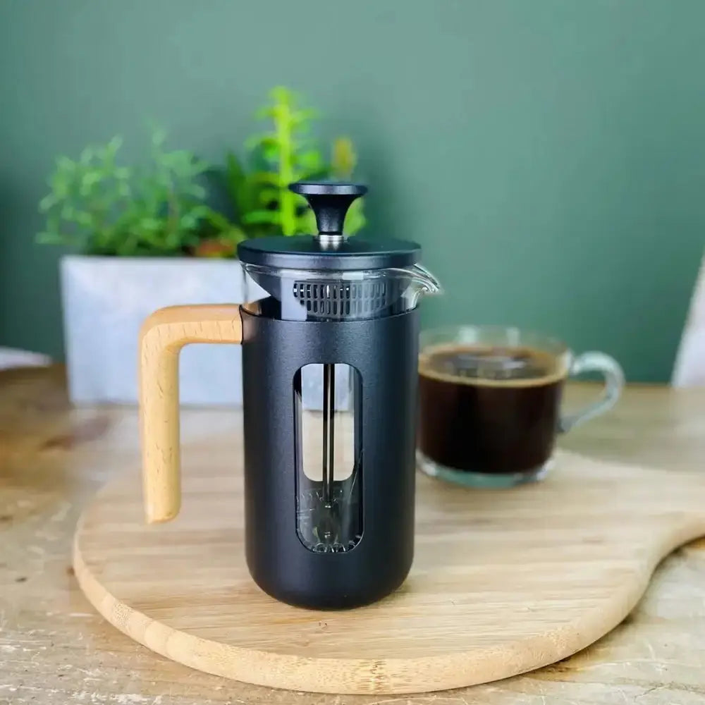 Black French press coffee maker with wooden handle on a wooden surface, with a blurred background of a plant and glass of coffee.