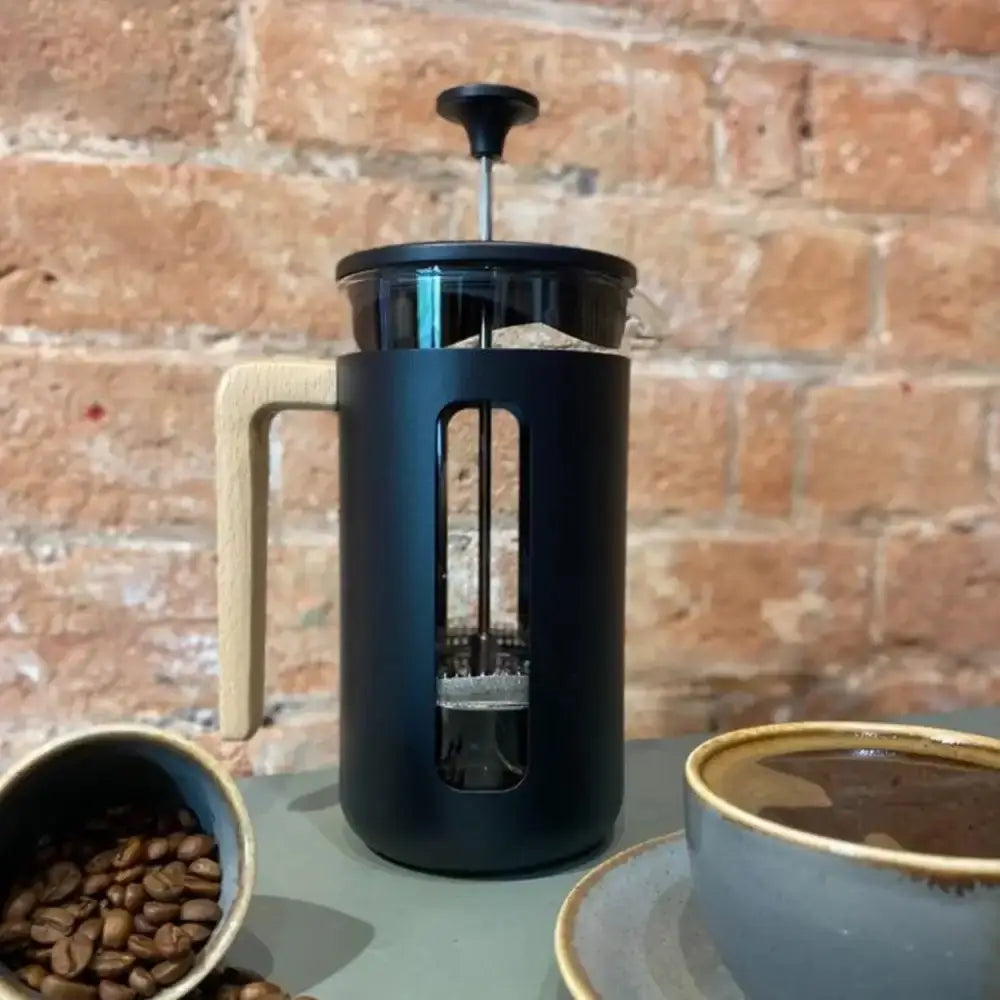 Black French press coffee maker with a bowl of coffee beans and a cup of coffee on a brick wall background.