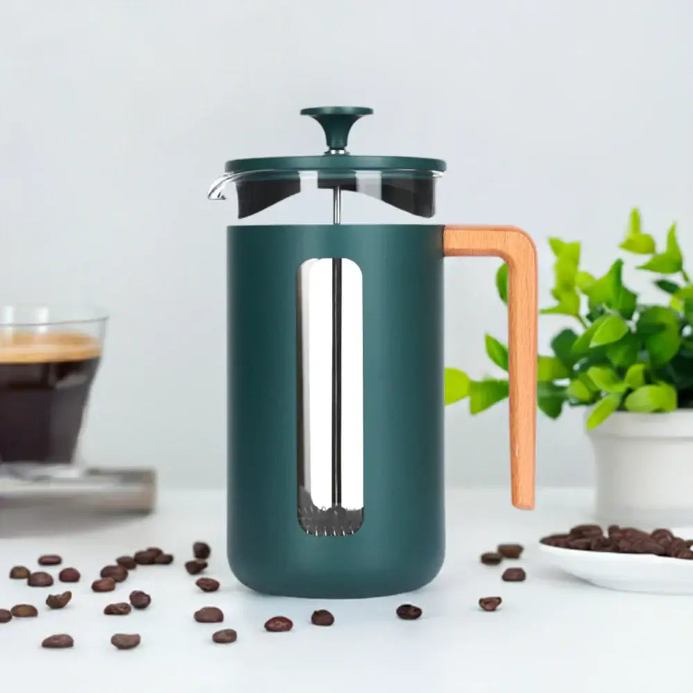 Green French press coffee maker with wooden handle on a white surface with coffee beans and a plant in the background.