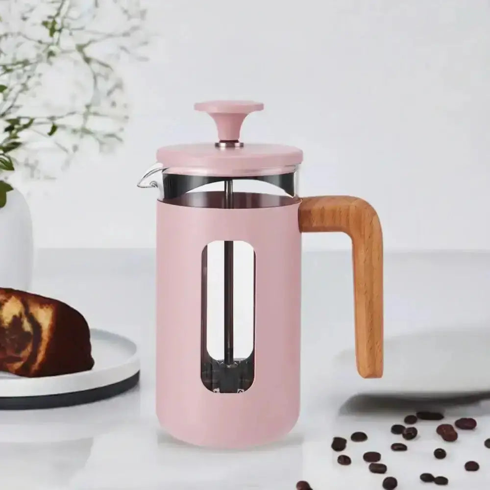 Pink French press coffee maker with wooden handle on a white surface with coffee beans and cake.
