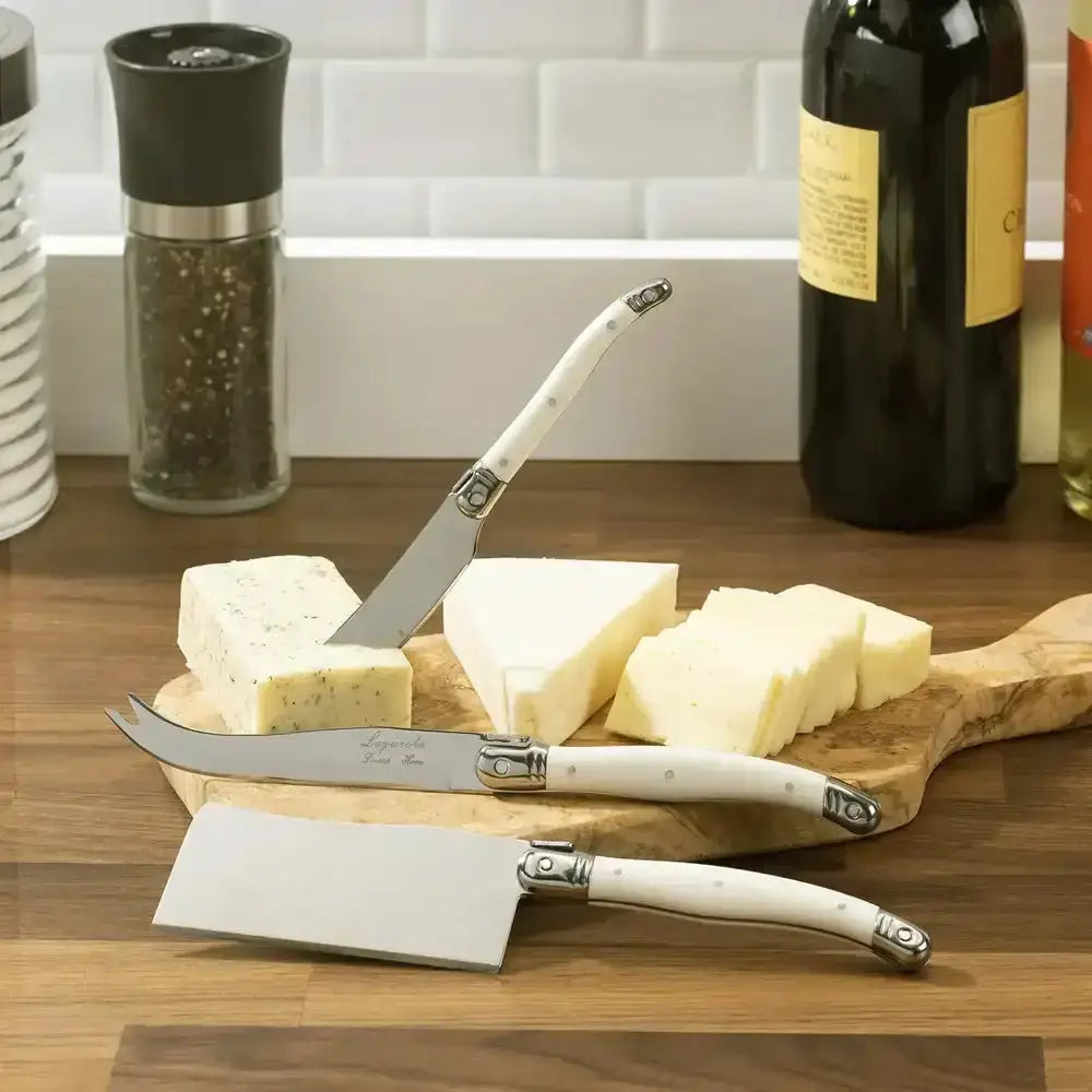 Three cheese knives with white handles on a wooden cutting board with cheese and wine bottles in the background.