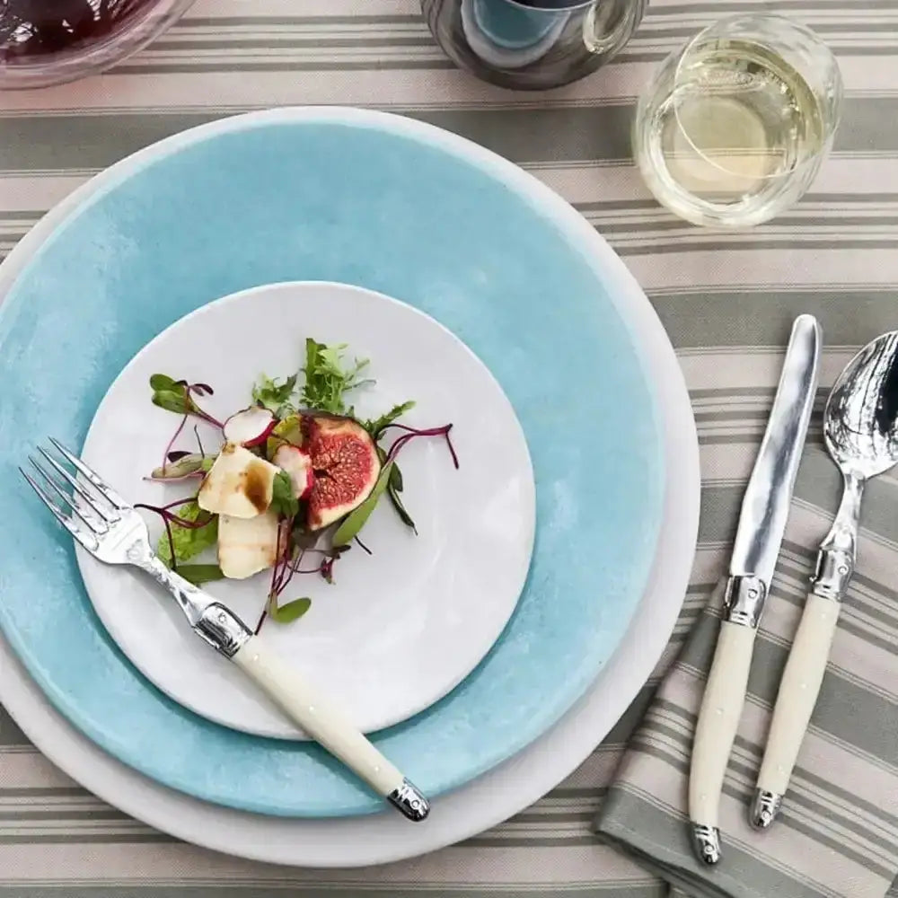 Dinner setting with a salad on a white plate, surrounded by silverware and glasses on a striped tablecloth.