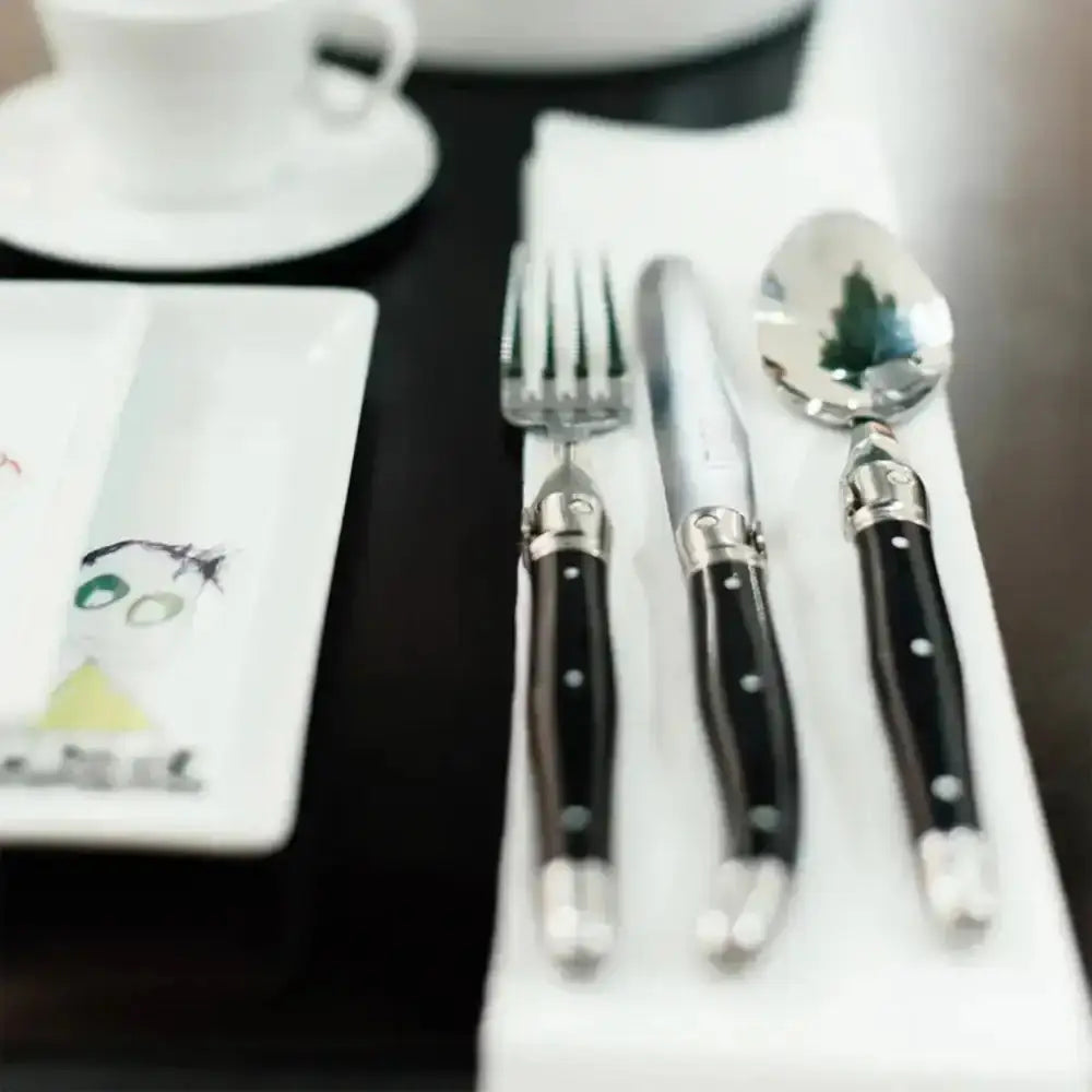 Silver cutlery set with black handles on a white surface, with a blurred background.