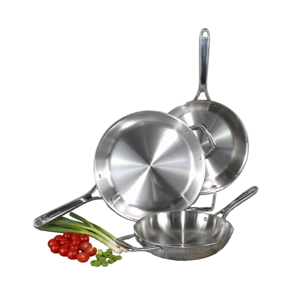 Set of stainless steel frying pans on a stone surface with vegetables in the foreground.