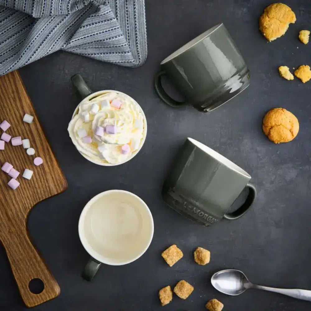 Four mugs on a dark surface with cookies, marshmallows, and a wooden board.