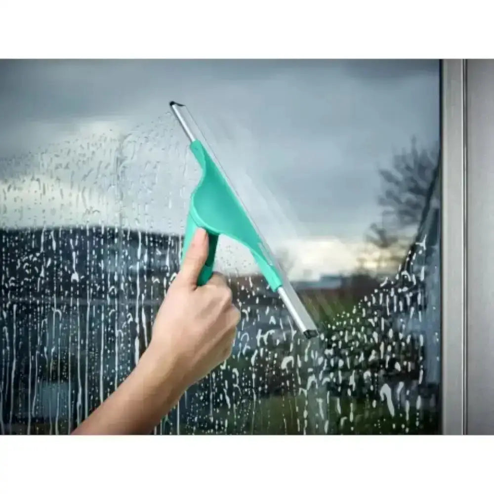 Hand using a green squeegee to clean a window with raindrops on a cloudy day.