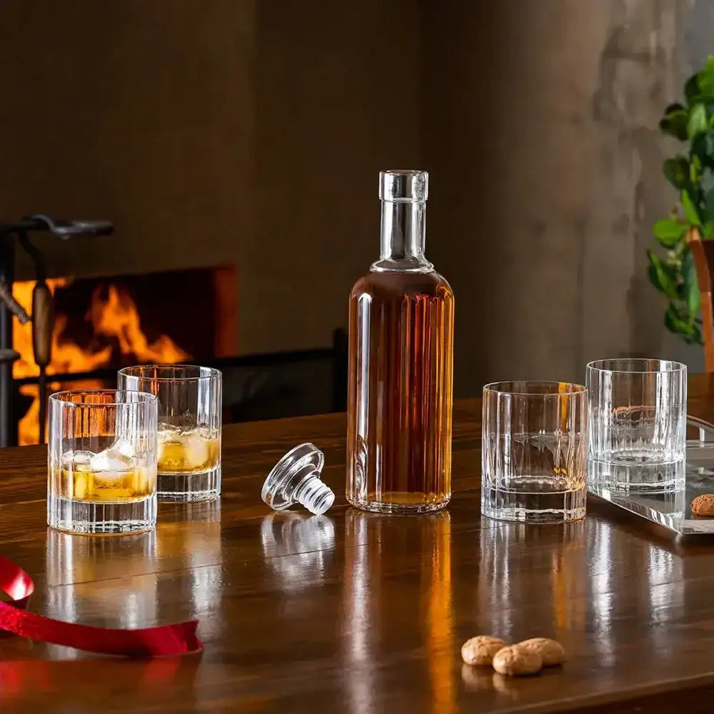 Bottle of whiskey with glasses on a wooden table in front of a fireplace.