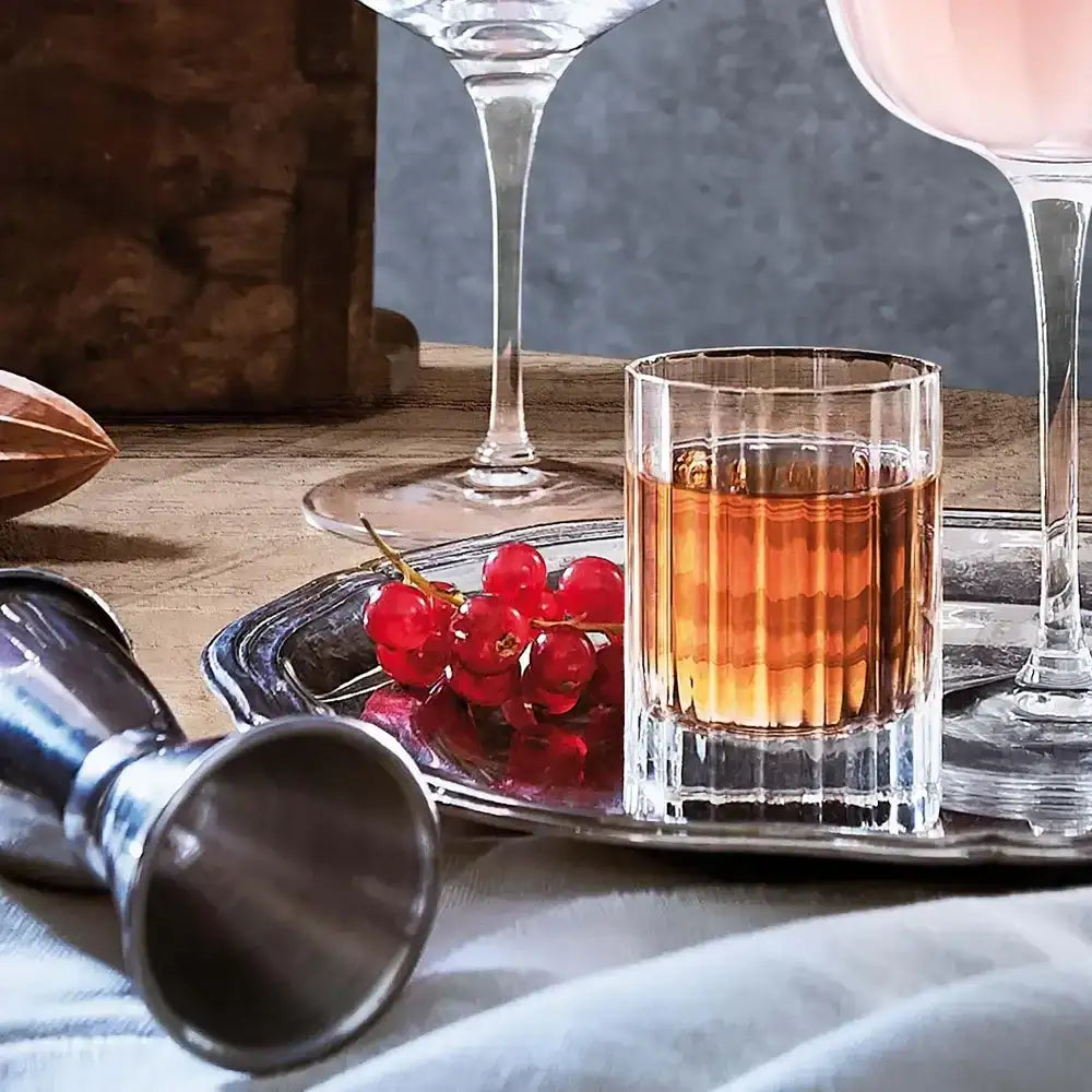 Glass of amber liquid with red berries on a tray, surrounded by cocktail-making tools.
