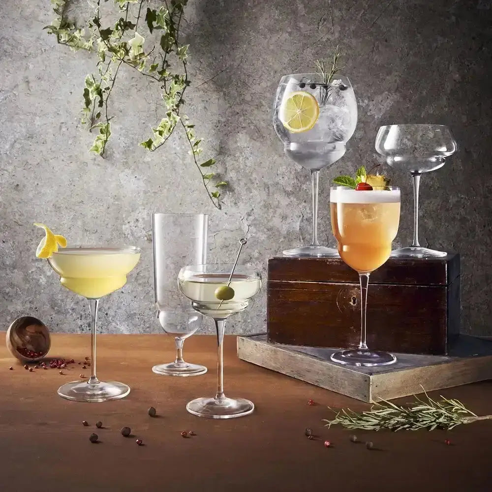 Assorted cocktails in various glasses on a wooden surface with a gray concrete wall background.