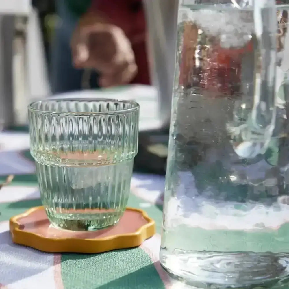 Clear glass with textured design on a yellow coaster next to a large clear pitcher on a checkered tablecloth.