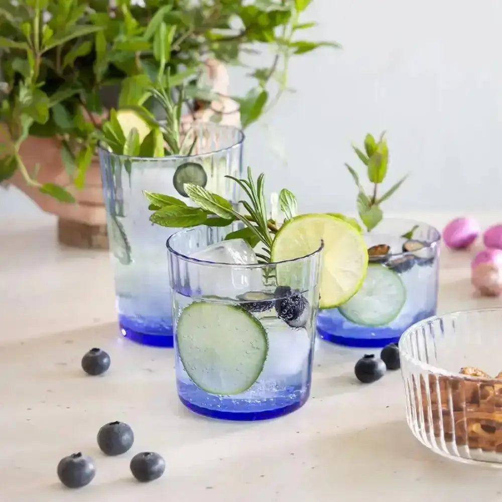 Three glasses of water with cucumber slices, lime, and blueberries on a wooden surface with plants in the background.