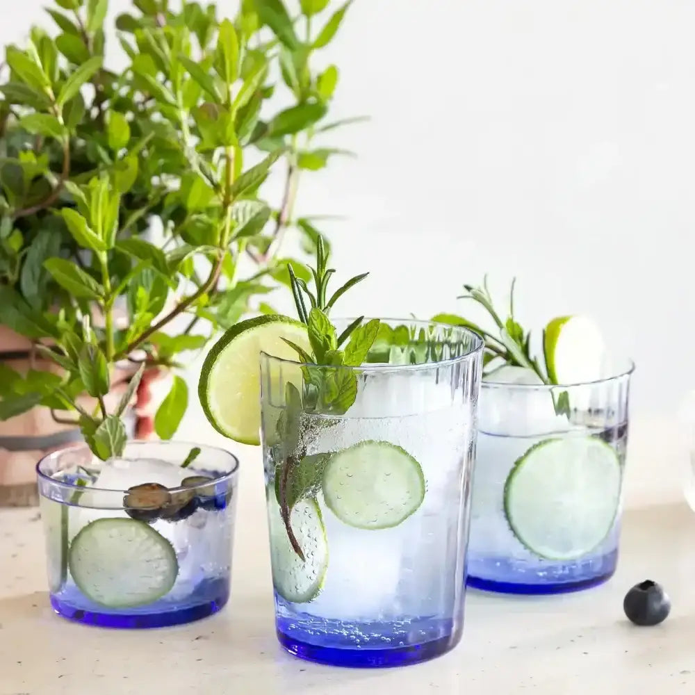 Three glasses of blue liquid with lime and mint on a white surface with plants in the background