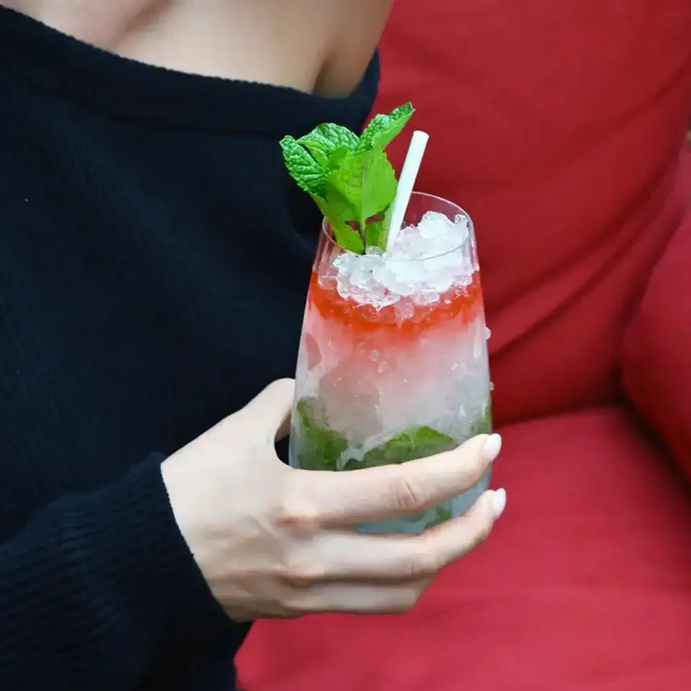 Hand holding a red and green cocktail with ice and mint leaves against a blurred background