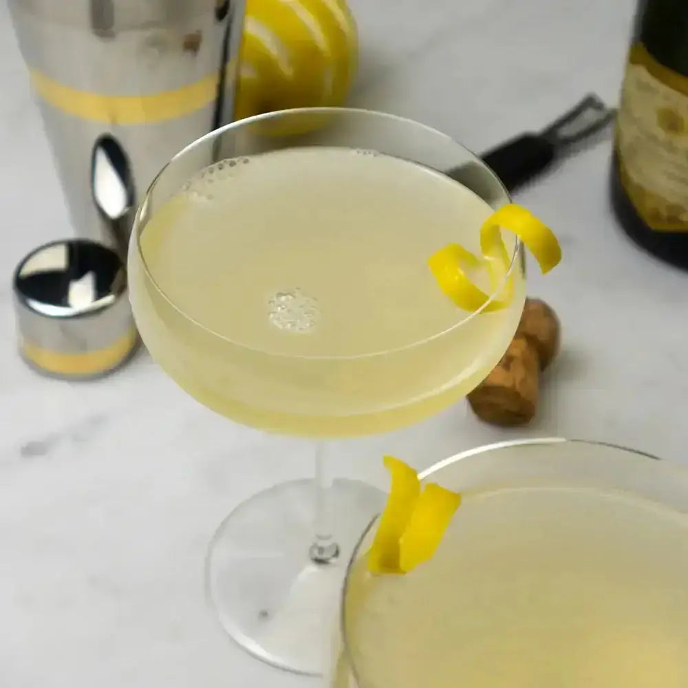 Two cocktails with lemon twists on a marble surface with bar tools in the background.