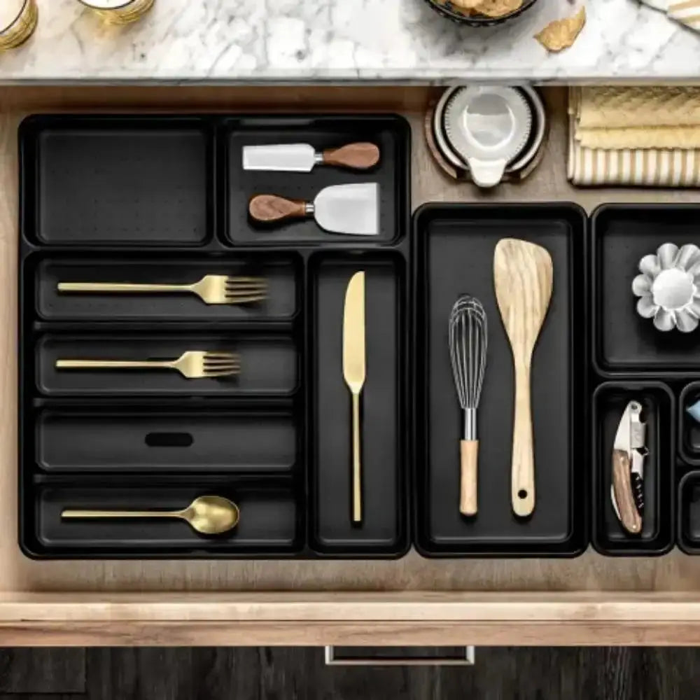 Kitchen drawer organizer with black compartments holding various utensils on a marble countertop.