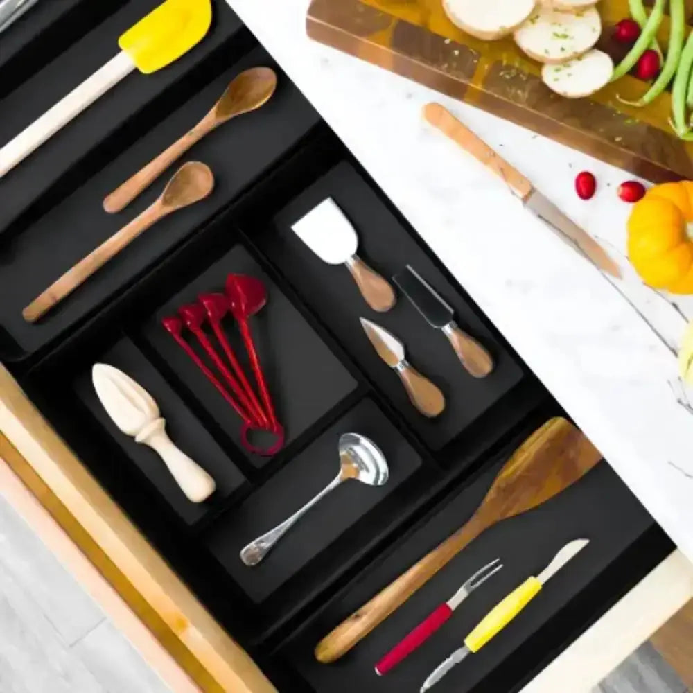 Set of kitchen utensils in a black drawer with a cutting board and vegetables in the background.