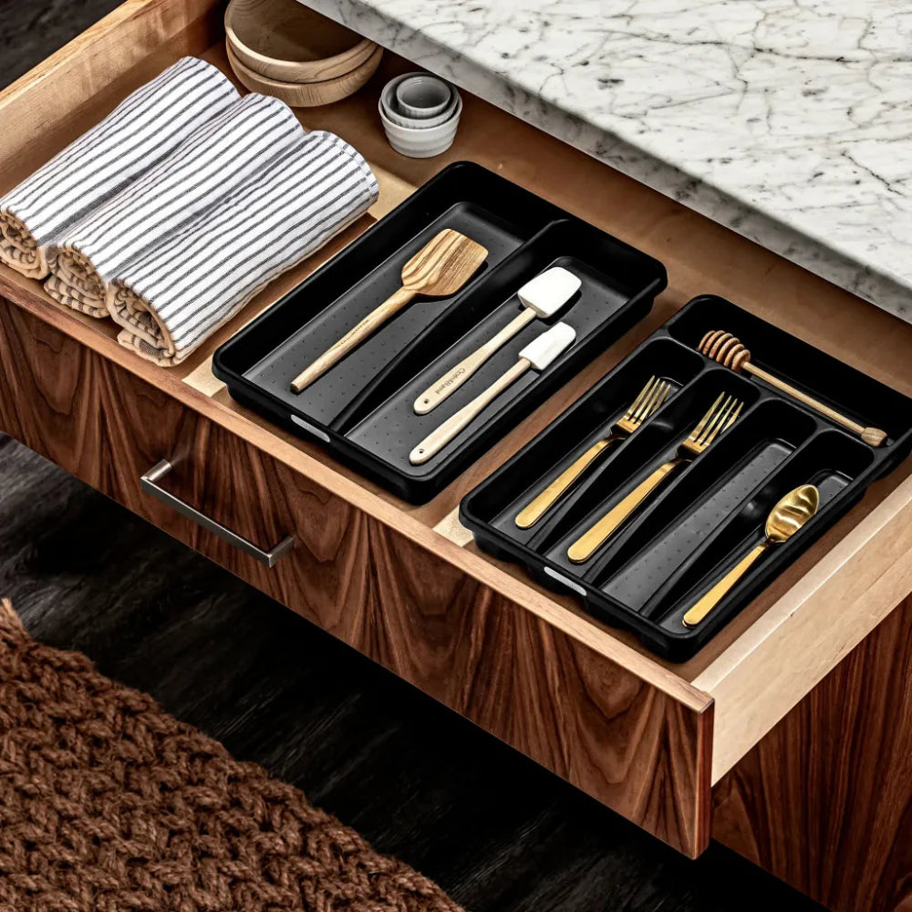 Wooden drawer with black trays containing kitchen utensils on a marble countertop.