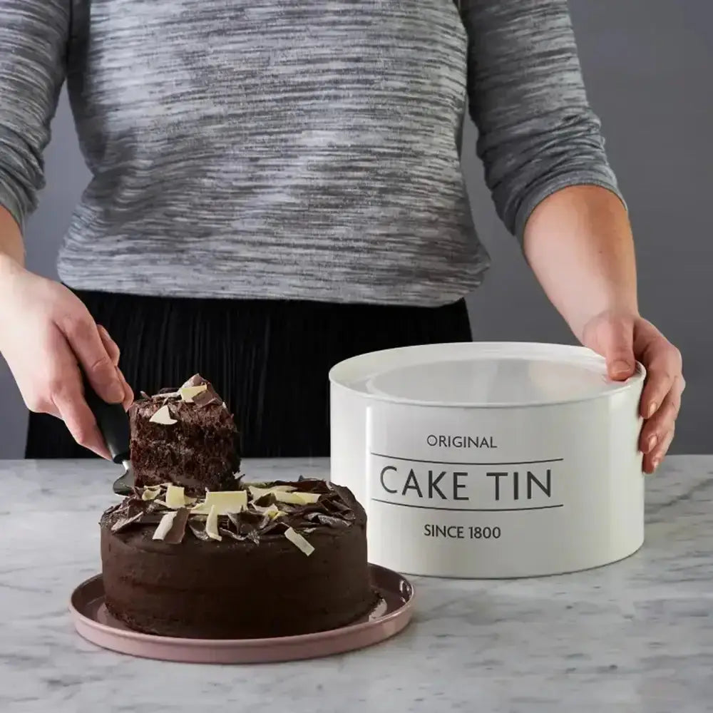Person cutting a chocolate cake next to a white cake tin labeled 'Original Cake Tin Since 1800' on a marble surface.