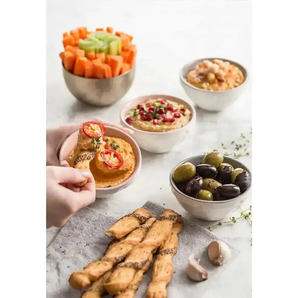 A hand holding a pink mason cash innovative mini food prep bowl filled with orange hummus topped with red peppers and herbs