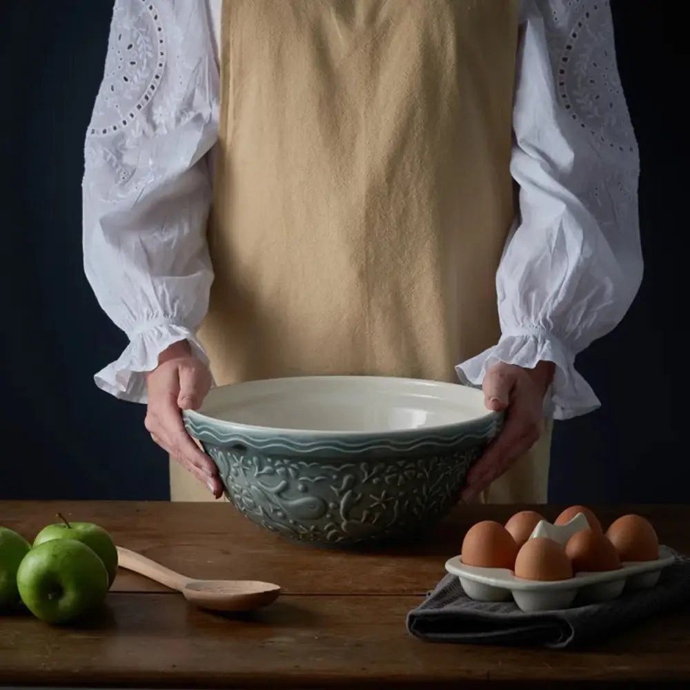 Person holding a green ceramic bowl on a wooden table with apples and eggs.