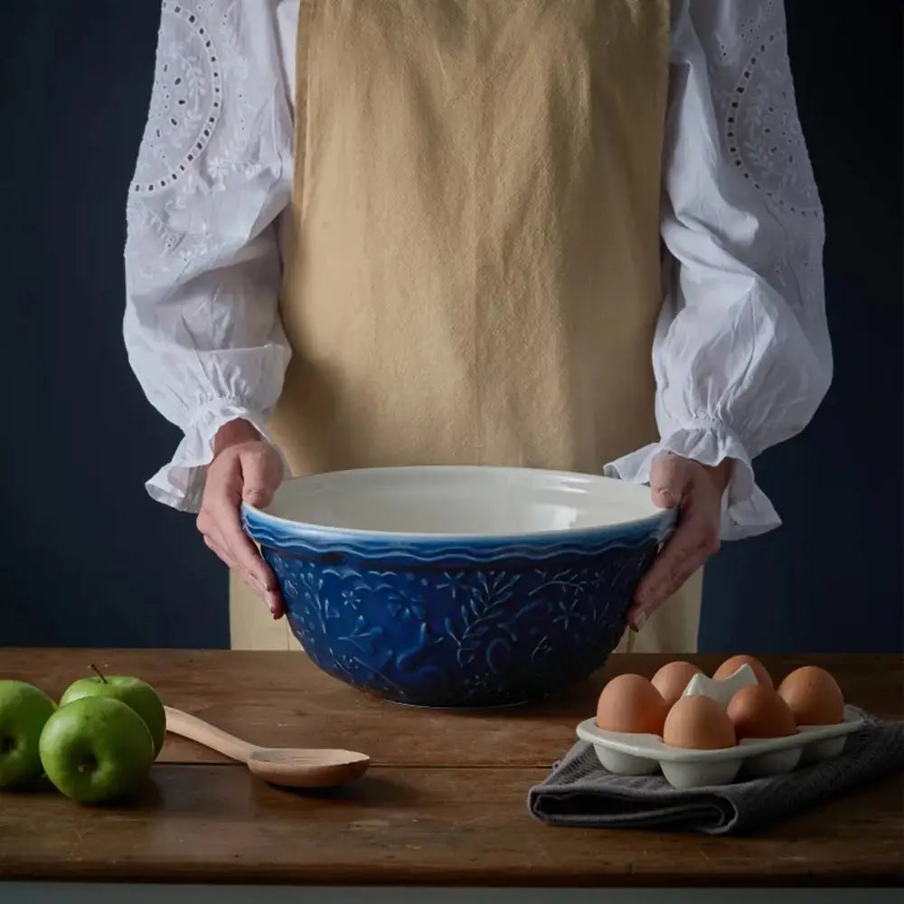 Person holding a blue ceramic bowl on a wooden table with apples and eggs.