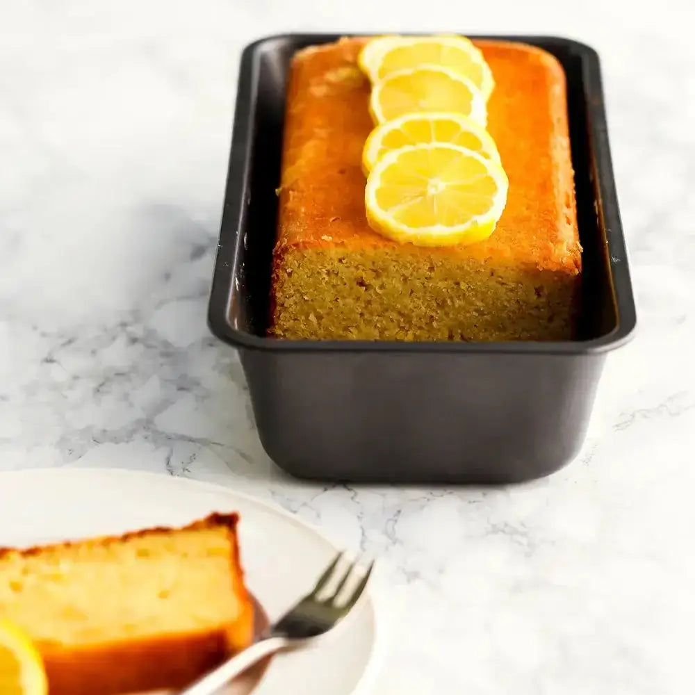 Loaf of lemon cake with slices on top, placed in a black loaf pan on a marble surface.