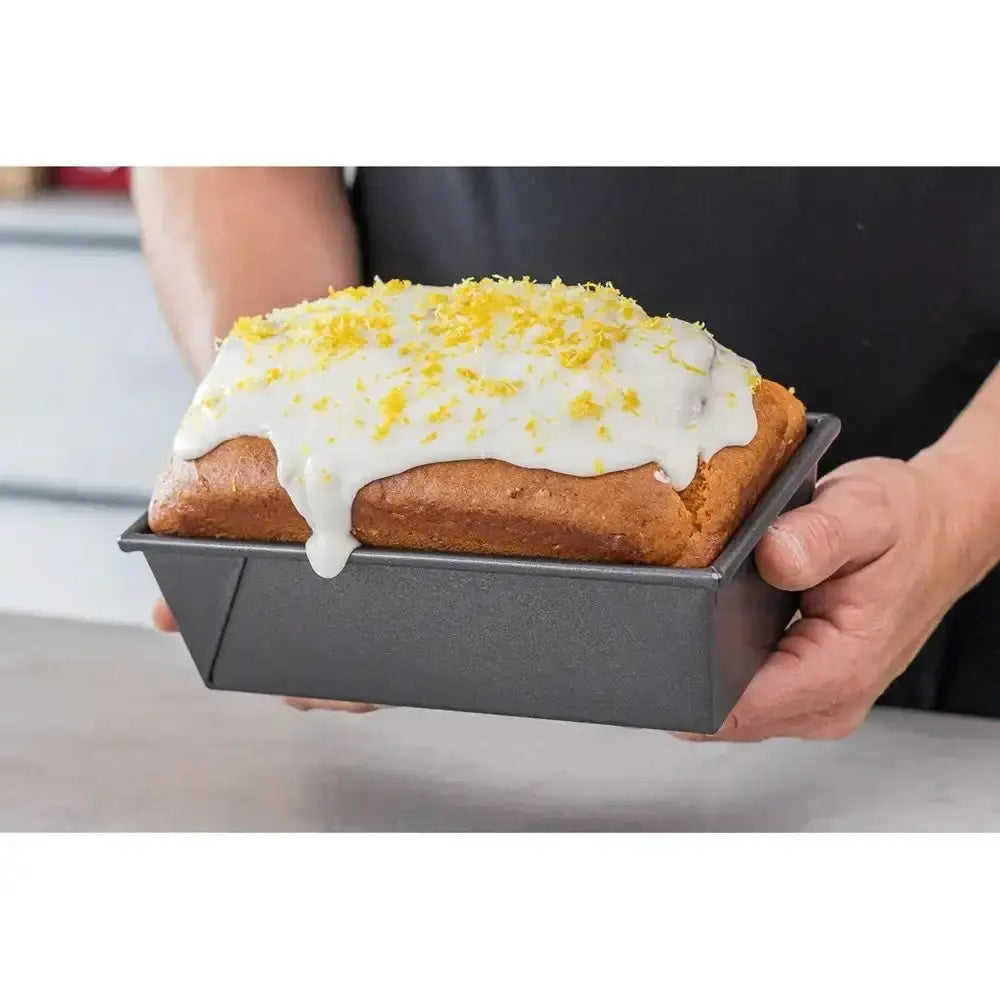 Person holding a loaf of bread with white icing and yellow sprinkles in a baking pan.